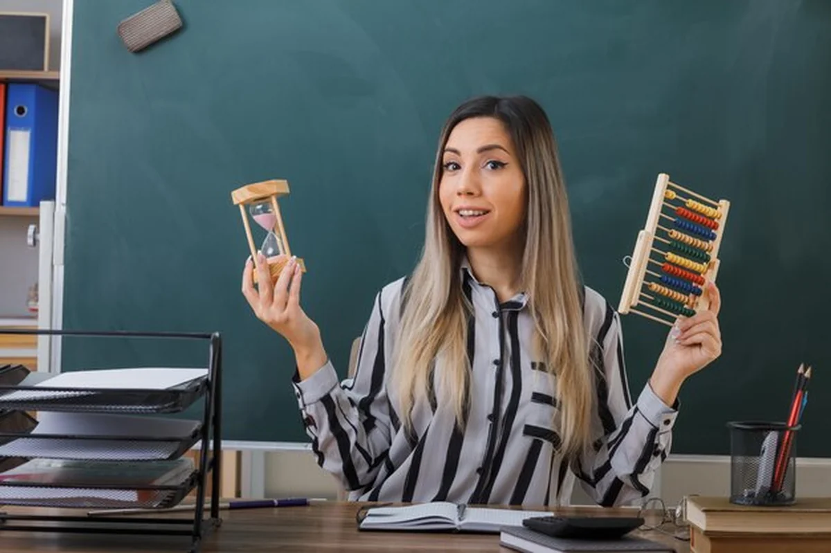 Nauczycielka przy tablicy, trzyma liczydło i klepsydrę, uśmiechnięta, w klasie